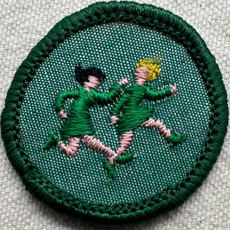 Puede incluir: Un parche redondo bordado en verde con dos figuras con vestidos verdes corriendo. Las figuras tienen piernas y brazos rosas, una con cabello negro y la otra rubio. El parche tiene un borde verde.