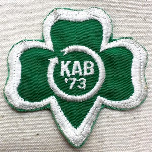 May include: Green and white embroidered patch in a cloverleaf shape. The patch features the text "KAB '73" inside a white circular design. The cloverleaf is outlined in white stitching.