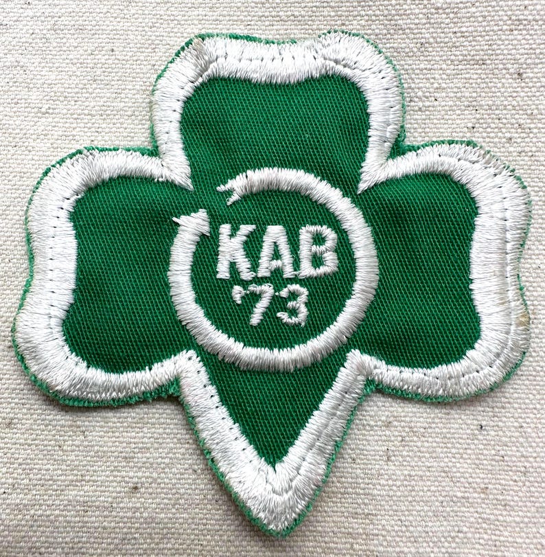 May include: Green and white embroidered patch in a cloverleaf shape. The patch features the text "KAB '73" inside a white circular design. The cloverleaf is outlined in white stitching.