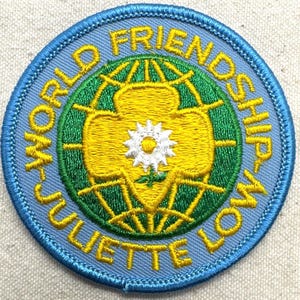 Peut inclure: Un &eacute;cusson rond avec une bordure bleu clair et le texte "WORLD FRIENDSHIP JULIETTE LOW". L'&eacute;cusson pr&eacute;sente un motif jaune et vert avec une fleur blanche au centre. Il est probablement destin&eacute; &agrave; une organisation de jeunesse.