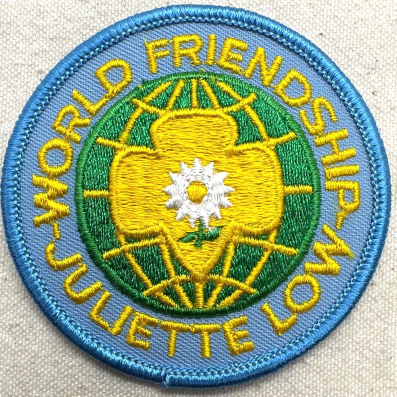 Peut inclure: Un &eacute;cusson rond avec une bordure bleu clair et le texte "WORLD FRIENDSHIP JULIETTE LOW". L'&eacute;cusson pr&eacute;sente un motif jaune et vert avec une fleur blanche au centre. Il est probablement destin&eacute; &agrave; une organisation de jeunesse.