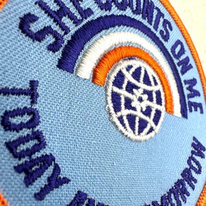 K&ouml;nnte beinhalten: Gestickter Aufn&auml;her mit dem Text "SHE COUNTS ON ME TODAY AND TOMORROW". Der Aufn&auml;her hat einen hellblauen Hintergrund, ein Regenbogen-Design und eine Globusgrafik. Der Rand ist orange.