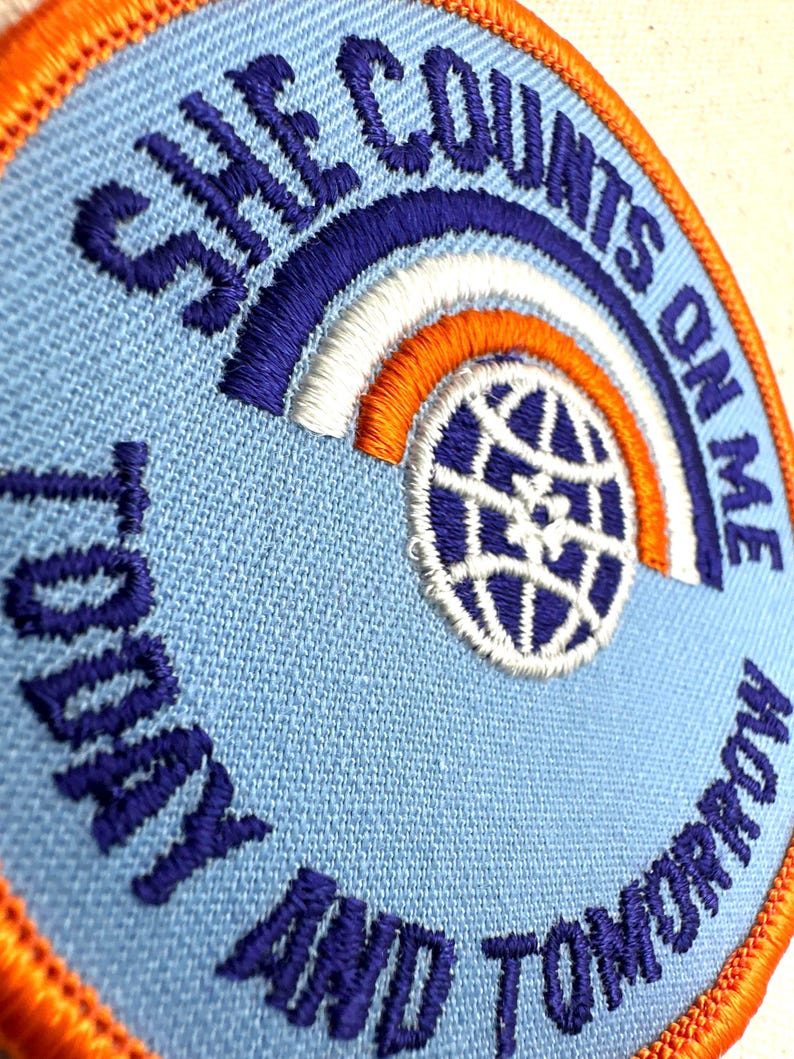 K&ouml;nnte beinhalten: Gestickter Aufn&auml;her mit dem Text "SHE COUNTS ON ME TODAY AND TOMORROW". Der Aufn&auml;her hat einen hellblauen Hintergrund, ein Regenbogen-Design und eine Globusgrafik. Der Rand ist orange.