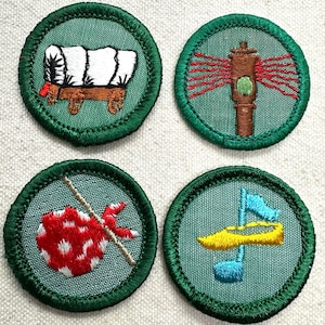 May include: Four embroidered patches with green borders. The patches feature a covered wagon, a tall brown structure with red lines, a red and white dotted sack on a stick, and a yellow and blue design. The patches are on a light beige background.