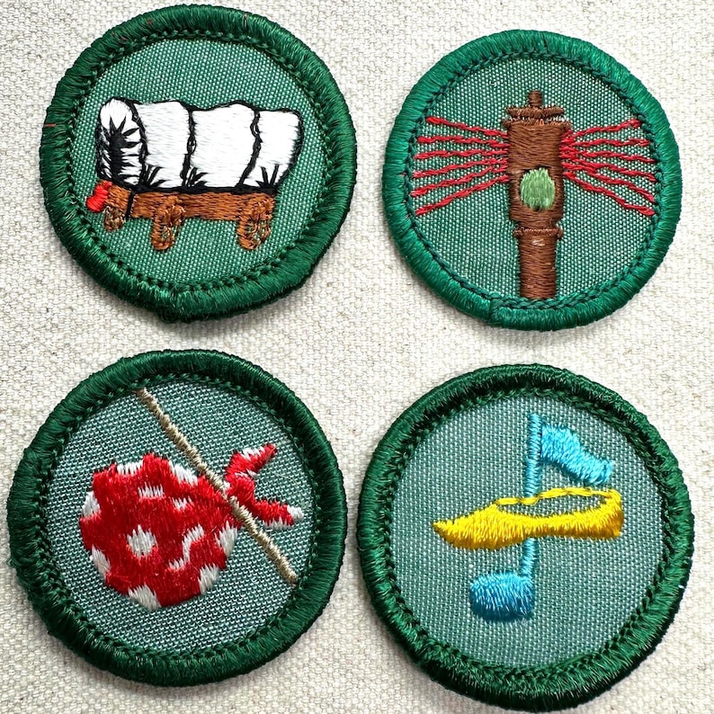 May include: Four embroidered patches with green borders. The patches feature a covered wagon, a tall brown structure with red lines, a red and white dotted sack on a stick, and a yellow and blue design. The patches are on a light beige background.
