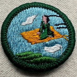 Puede incluir: Parche bordado que representa a una persona en una alfombra voladora sobre un cielo azul con nubes blancas. La persona lleva ropa verde y rosa. El parche tiene un borde verde y una alfombra amarilla.
