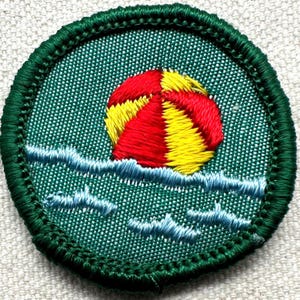 Puede incluir: Parche bordado con una pelota de playa roja y amarilla flotando sobre olas azules. El parche tiene un borde verde y un fondo verde texturizado. Un dise&ntilde;o divertido y colorido.
