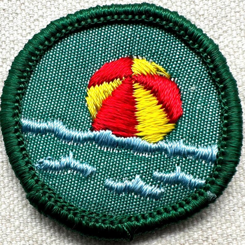 Puede incluir: Parche bordado con una pelota de playa roja y amarilla flotando sobre olas azules. El parche tiene un borde verde y un fondo verde texturizado. Un dise&ntilde;o divertido y colorido.
