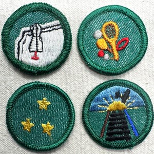 May include: Four embroidered patches with green borders and backgrounds. The patches feature a bell, tennis equipment, three gold stars, and a stylized landscape with a sun and a structure. The patches are circular.