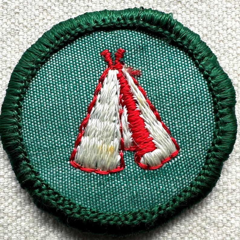 May include: Embroidered patch featuring a red and white teepee design on a green background. The teepee is outlined in red thread, and the patch has a green stitched border. This patch is likely for a scout or outdoor activity.