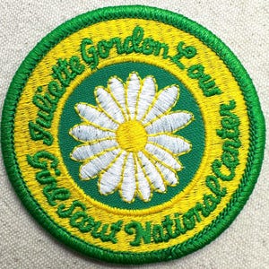 Puede incluir: Parche bordado con borde verde y centro amarillo y verde. El centro presenta una margarita blanca con centro amarillo. El texto bordado en amarillo dice "Juliette Gordon Low" y "Girl Scout National Center".