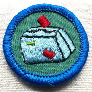 May include: Embroidered patch with a blue border and a light green center. The patch features a light blue object with black and red accents. The object has a red rectangle on top and a red heart shape on the bottom. The patch is round.