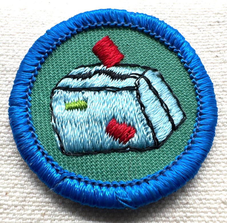 May include: Embroidered patch with a blue border and a light green center. The patch features a light blue object with black and red accents. The object has a red rectangle on top and a red heart shape on the bottom. The patch is round.