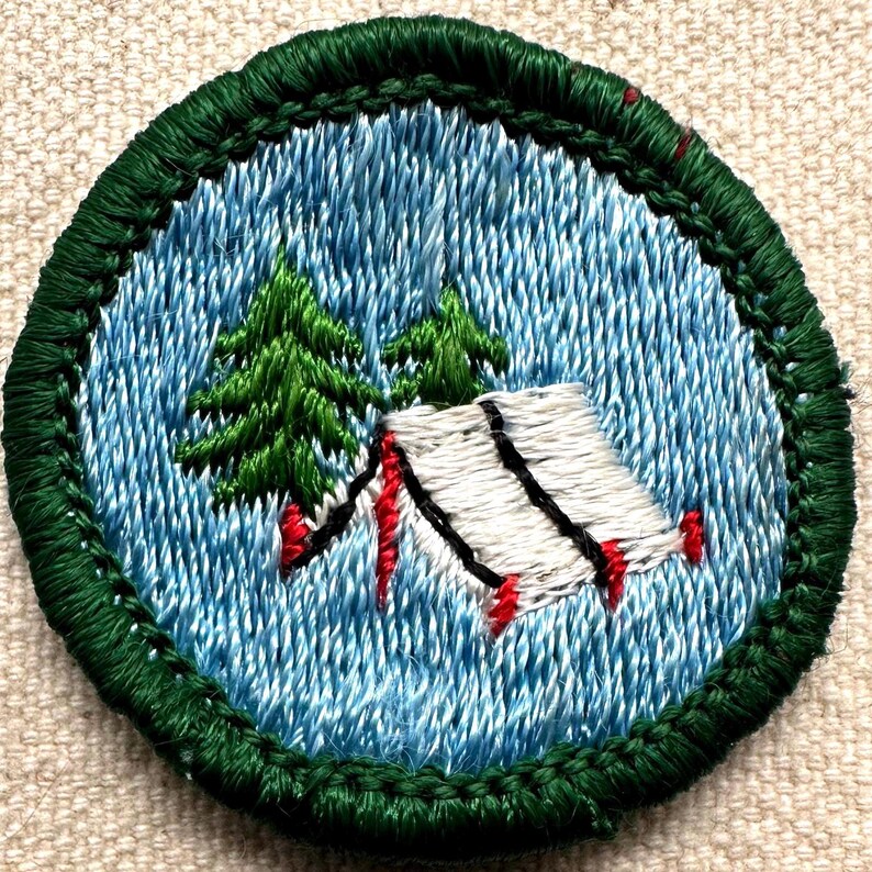 Puede incluir: Parche bordado con una escena de acampada. El parche tiene un borde verde y un fondo azul claro. Se representa una tienda blanca con l&iacute;neas negras y detalles rojos, junto con dos pinos verdes. El parche es redondo.