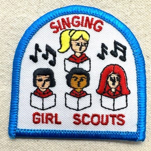 May include: Embroidered patch with a blue border and the words "SINGING GIRL SCOUTS". The patch features illustrations of three girls singing from books, with musical notes on either side. The girls have blonde, black, and red hair.