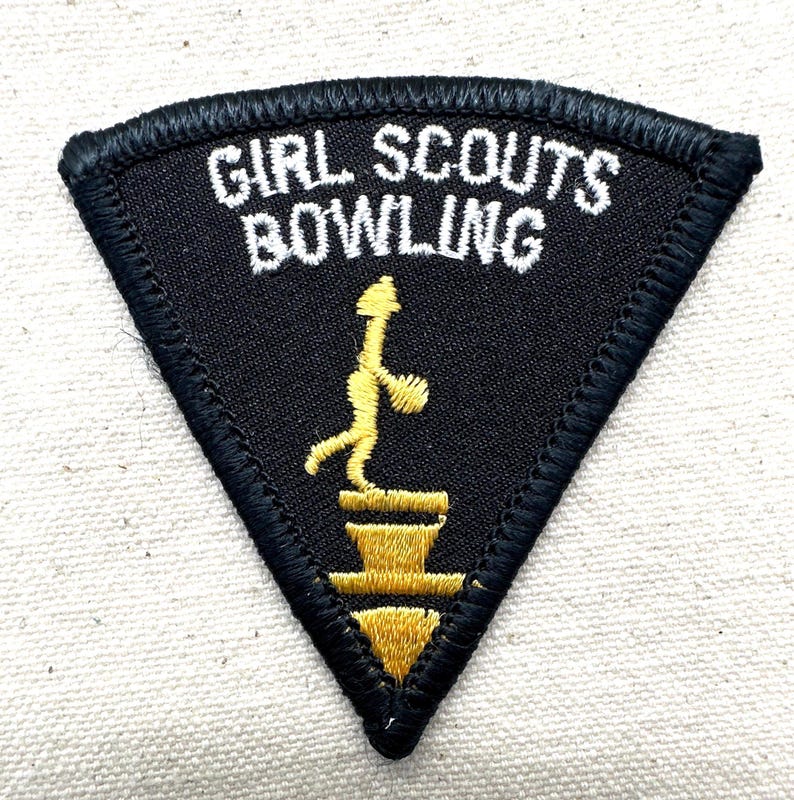 Puede incluir: Parche triangular negro con texto bordado blanco "GIRL SCOUTS BOWLING". Una figura amarilla de un jugador de bolos est&aacute; sobre un pedestal amarillo. El parche tiene un borde negro.