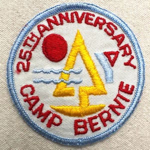 May include: Embroidered patch with a blue border and the text "25th Anniversary Camp Bernie" in red. The patch features a red sun, yellow sailboat, blue waves, and a blue and red triangle.