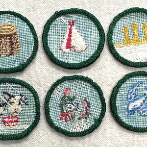 May include: Six embroidered patches with green borders and various designs. The designs include a basket, a teepee, three yellow structures, a cow drinking from a bowl, flowers, and a blue bird.