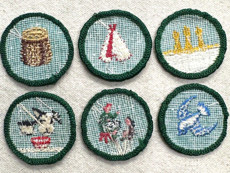 May include: Six embroidered patches with green borders and various designs. The designs include a basket, a teepee, three yellow structures, a cow drinking from a bowl, flowers, and a blue bird.