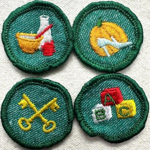 May include: Four circular embroidered patches with green borders. The patches feature various designs: a mortar and pestle, a pumpkin with a plane, crossed keys, and alphabet blocks. The patches are on a textured, off-white background.