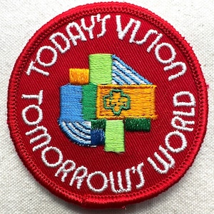 May include: A round, red embroidered patch with the words "TODAY'S VISION WORLD TOMORROW'S" in white. The center features a colorful design with blue, green, yellow, and orange elements. A cross-like shape is in the center.