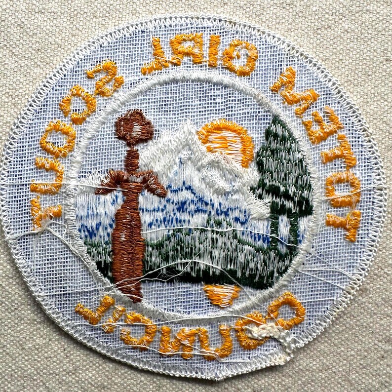 K&ouml;nnte beinhalten: Runder, gestickter Aufn&auml;her mit einer Berglandschaft, einem Schwert und Text. Der Aufn&auml;her hat einen wei&szlig;en Hintergrund mit einer blau-gr&uuml;nen Landschaft, einem braunen Schwert und orangefarbenem Text mit der Aufschrift "Golden Spirit Canada".