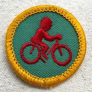 Puede incluir: Parche bordado circular con un ciclista rojo en bicicleta sobre un fondo verde. El parche tiene un borde amarillo. El dise&ntilde;o es una representaci&oacute;n estilizada de una persona montando en bicicleta.
