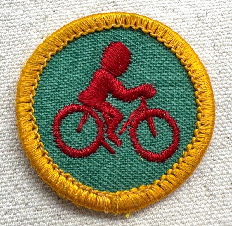 Puede incluir: Parche bordado circular con un ciclista rojo en bicicleta sobre un fondo verde. El parche tiene un borde amarillo. El dise&ntilde;o es una representaci&oacute;n estilizada de una persona montando en bicicleta.