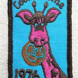 May include: Embroidered patch featuring a giraffe holding a cookie. The giraffe is pink and brown, set against a blue background. The text "Cookie Time" and "1976" are displayed on the patch, which has a brown border.