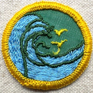May include: Embroidered patch featuring a stylized ocean wave design in shades of blue and green, with two yellow birds in flight. The circular patch has a yellow border, and is likely for a jacket or bag.
