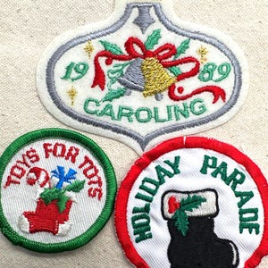 May include: Three embroidered Christmas patches. One is an ornament shape with a bell, ribbon, and the text "CAROLING 1989". The other two are round, with the text "TOYS FOR TOTS" and "HOLIDAY PARADE" with festive imagery.