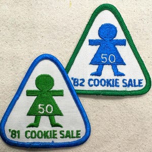 May include: Two triangular patches with a white background, featuring a blue and green outline. Each patch has a stylized figure with outstretched arms and legs, the number "50", and the text "COOKIE SALE" with the years "'81" and "'82".