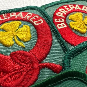 May include: Close-up of several embroidered patches. Each patch features a yellow shamrock within a red circle, with the words "BE PREPARED" in white. The patches have a green background and are edged with green stitching.