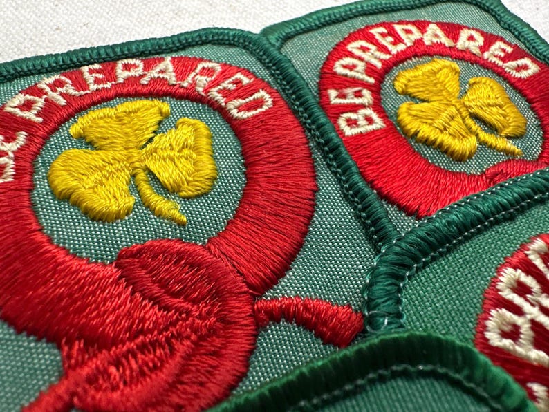 May include: Close-up of several embroidered patches. Each patch features a yellow shamrock within a red circle, with the words "BE PREPARED" in white. The patches have a green background and are edged with green stitching.