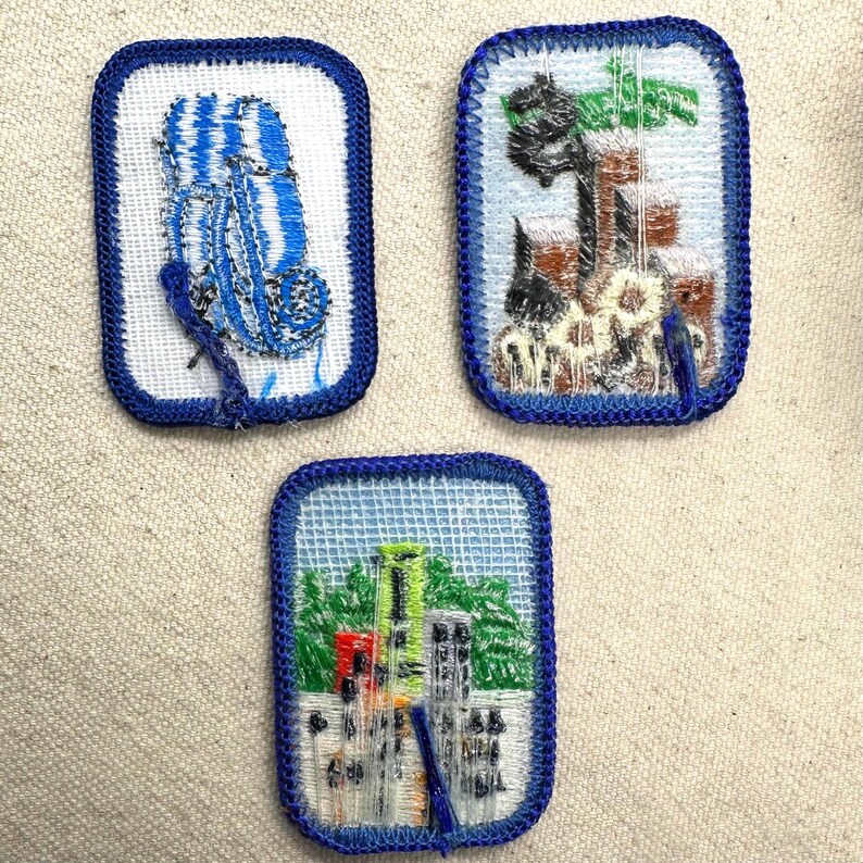 May include: Three embroidered patches with blue borders. One patch depicts a blue and white striped object. Another shows a landscape with a tree and flowers. The third patch features a cityscape with green and yellow buildings.
