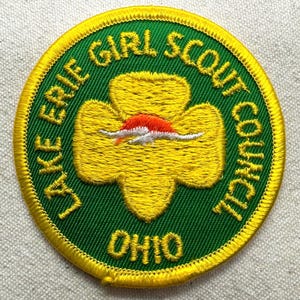 May include: A circular embroidered patch with a green background and a yellow border. The text reads Lake Erie Girl Scout Council Ohio. In the center is a yellow trefoil shape featuring a stylized orange and white graphic of a rising sun.