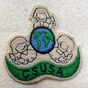 K&ouml;nnte beinhalten: Gestickter Aufn&auml;her mit einem Globus mit blauen Ozeanen und gr&uuml;nen Kontinenten, umgeben von drei stilisierten Figuren. Der Aufn&auml;her hat ein gr&uuml;nes Banner mit der Aufschrift "GSUSA". Der Aufn&auml;her ist beige mit schwarzen Umrissen.