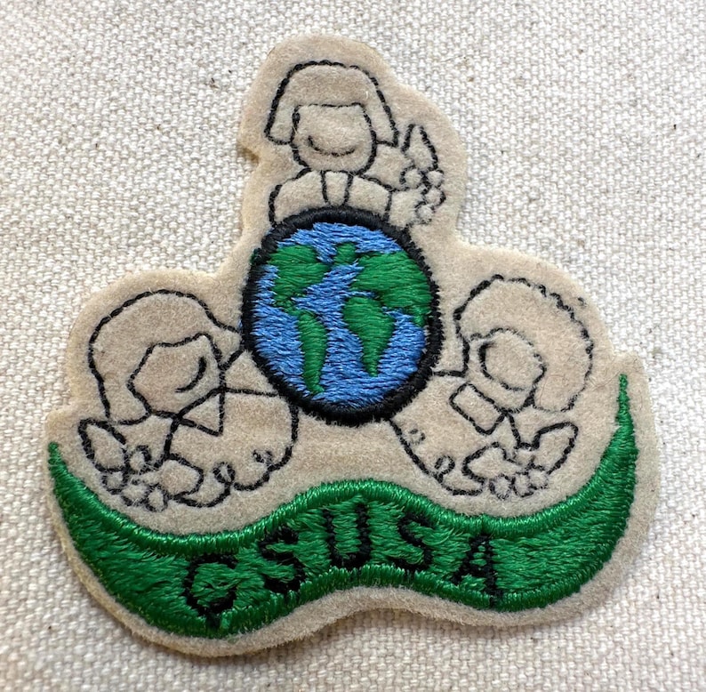 K&ouml;nnte beinhalten: Gestickter Aufn&auml;her mit einem Globus mit blauen Ozeanen und gr&uuml;nen Kontinenten, umgeben von drei stilisierten Figuren. Der Aufn&auml;her hat ein gr&uuml;nes Banner mit der Aufschrift "GSUSA". Der Aufn&auml;her ist beige mit schwarzen Umrissen.