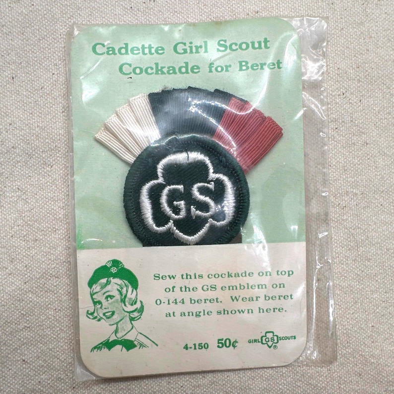 May include: Vintage Cadette Girl Scout cockade for beret, packaged in clear plastic. The package features a green emblem with white stitching and the letters "GS". The cockade has white, black, and red ribbons. Text on the package reads "Cadette Girl Scout Cockade for Beret."
