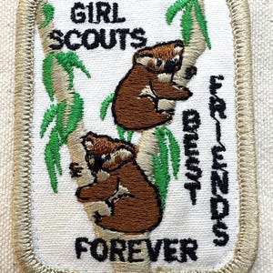 K&ouml;nnte beinhalten: Gestickter Girl Scouts-Aufn&auml;her mit zwei braunen Koalas auf einem Baum mit gr&uuml;nen Bl&auml;ttern. Der Aufn&auml;her tr&auml;gt den schwarzen Schriftzug "GIRL SCOUTS BEST FRIENDS FOREVER" mit einem beigen Rand.