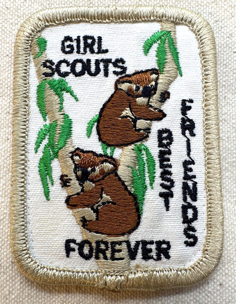 K&ouml;nnte beinhalten: Gestickter Girl Scouts-Aufn&auml;her mit zwei braunen Koalas auf einem Baum mit gr&uuml;nen Bl&auml;ttern. Der Aufn&auml;her tr&auml;gt den schwarzen Schriftzug "GIRL SCOUTS BEST FRIENDS FOREVER" mit einem beigen Rand.