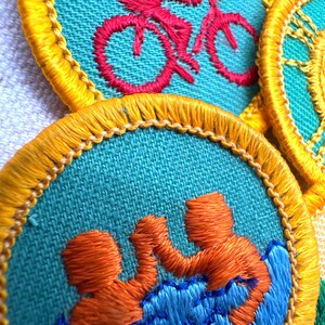Puede incluir: Primer plano de parches bordados con tela turquesa y bordes amarillos. Un parche presenta un ciclista rojo, otro muestra dos figuras naranjas en agua azul. Los parches son probablemente para una organizaci&oacute;n juvenil.