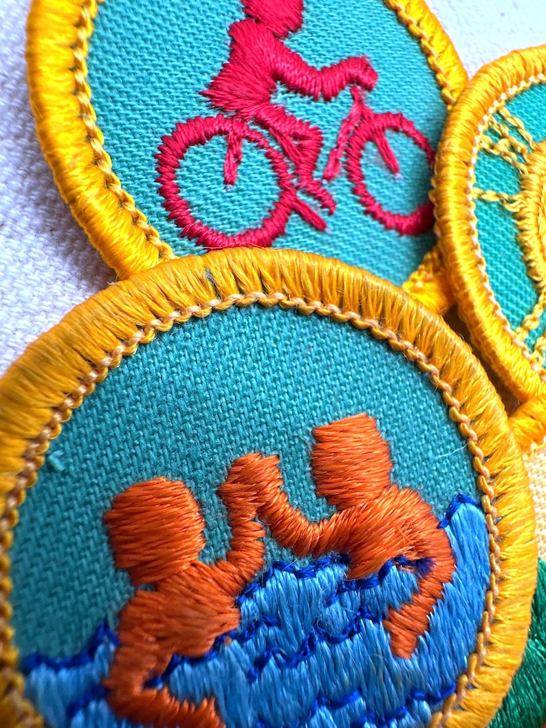 Puede incluir: Primer plano de parches bordados con tela turquesa y bordes amarillos. Un parche presenta un ciclista rojo, otro muestra dos figuras naranjas en agua azul. Los parches son probablemente para una organizaci&oacute;n juvenil.