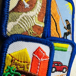 May include: A collection of embroidered patches with blue borders. One patch depicts a climber on a rock face, another shows a red car on a road, and others feature buildings and abstract designs. The patches are colorful and detailed.