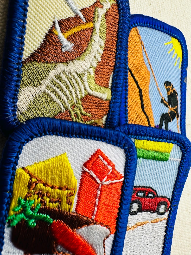 May include: A collection of embroidered patches with blue borders. One patch depicts a climber on a rock face, another shows a red car on a road, and others feature buildings and abstract designs. The patches are colorful and detailed.