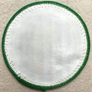 Op de afbeelding: Een rond, wit object met een groene gehaakte rand. Het witte midden heeft een licht getextureerd uiterlijk. De groene rand is gemaakt van een strak gehaakt patroon. Het object staat tegen een neutrale, getextureerde achtergrond.