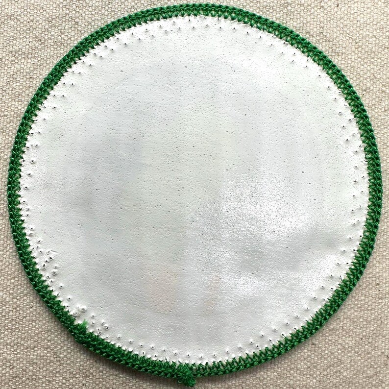 Op de afbeelding: Een rond, wit object met een groene gehaakte rand. Het witte midden heeft een licht getextureerd uiterlijk. De groene rand is gemaakt van een strak gehaakt patroon. Het object staat tegen een neutrale, getextureerde achtergrond.