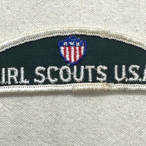 May include: A vintage Girl Scouts U.S.A. patch. The patch is dark green with white embroidered text and a shield emblem featuring red and white stripes and blue stars. The patch has a white border.