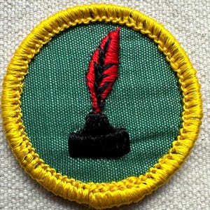 Pode incluir: Patch bordado com fundo verde e borda amarela. O patch apresenta uma pena vermelha com detalhes pretos sobre um tinteiro preto. O patch &eacute; circular.