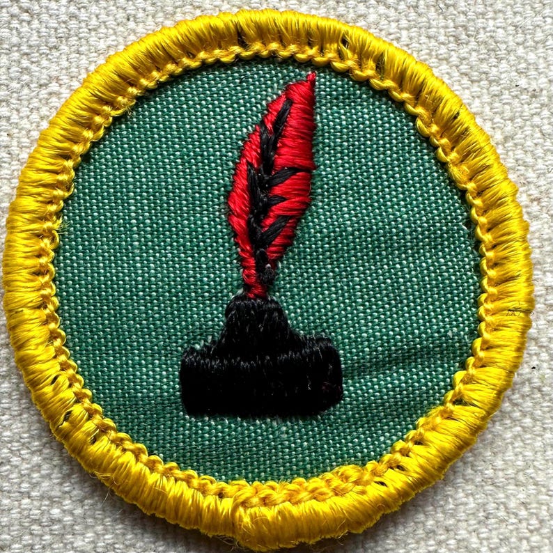 Pode incluir: Patch bordado com fundo verde e borda amarela. O patch apresenta uma pena vermelha com detalhes pretos sobre um tinteiro preto. O patch &eacute; circular.